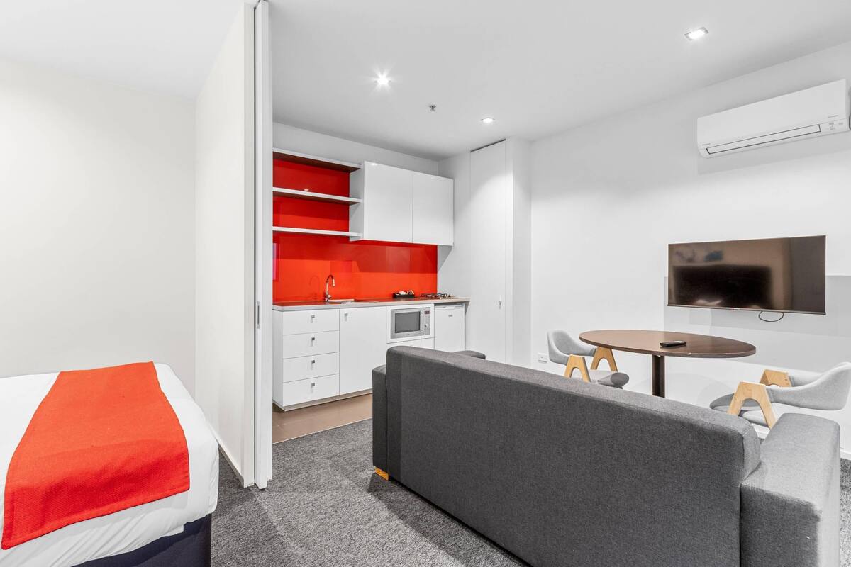 Una habitación de hotel moderna con cama, sofá gris, mesa de comedor y kitchenette.
