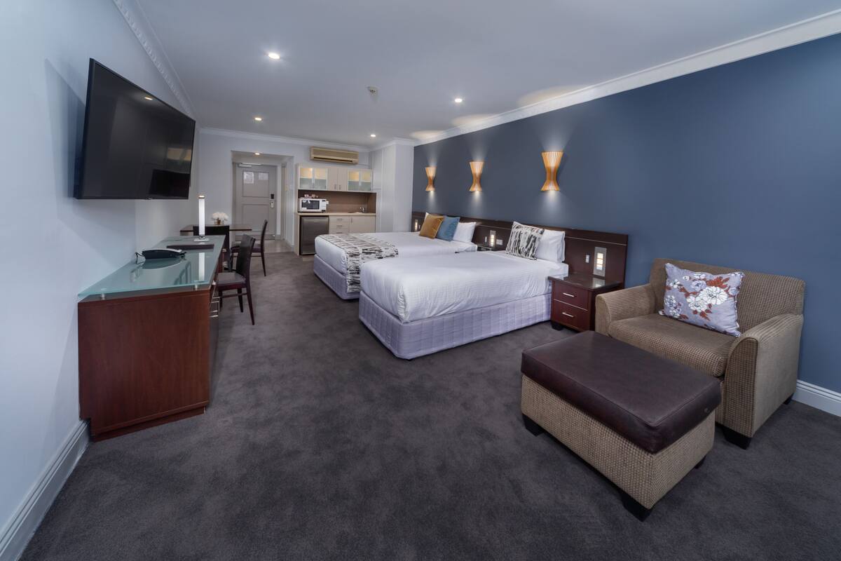 A hotel room with a large bed, a sofa, a desk, and a TV. There is a kitchenette in the background.