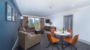 Habitación de hotel con zona de comedor, sofá, televisión y balcón con vistas a la ciudad.