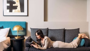 A woman lying on a dark sofa, reading a book, with a gold lamp on a side table and a framed abstract artwork on the wall. - The Capitol Hotel Sydney (Haymarket)