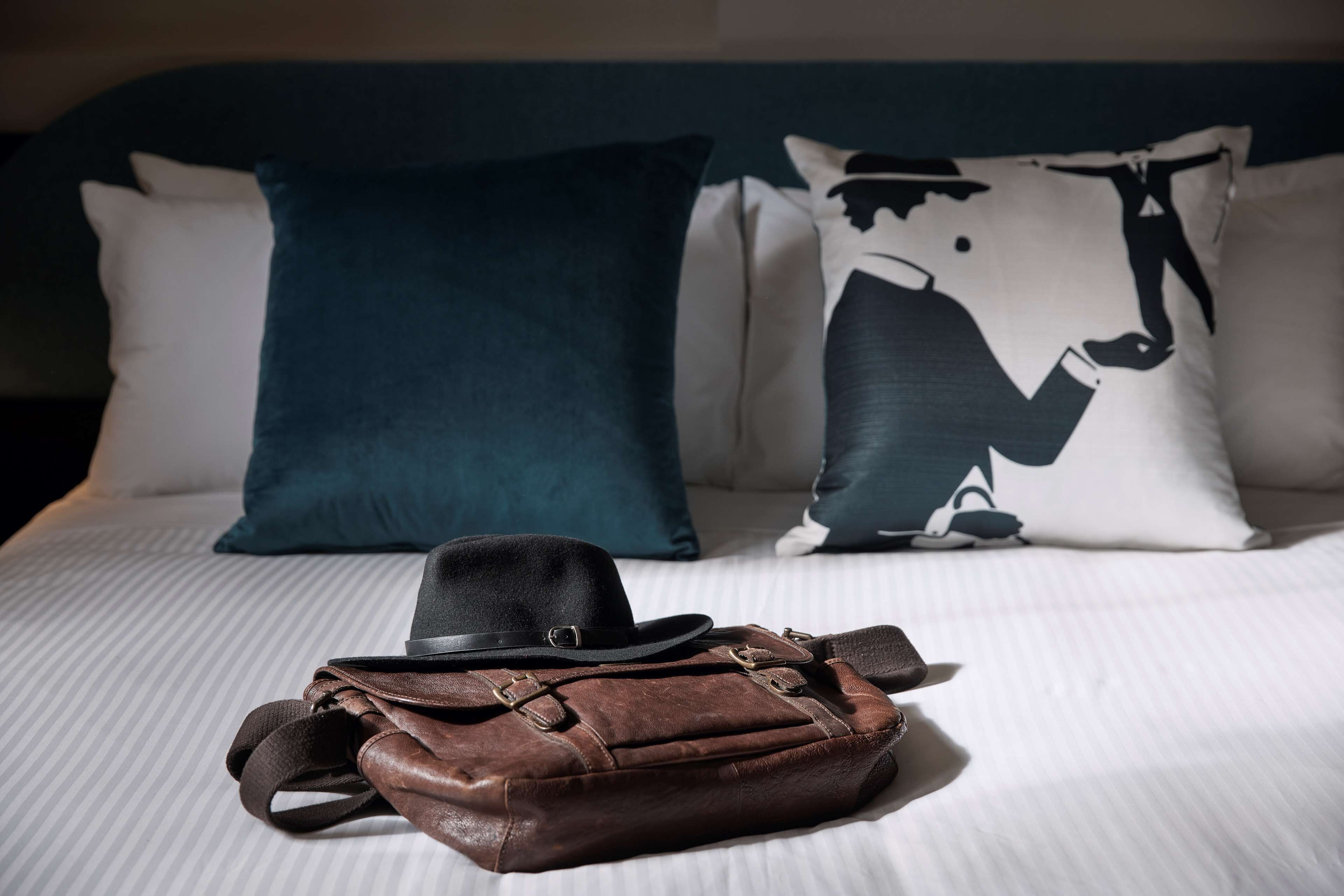 A bed with pillows and a brown leather bag and hat on it.