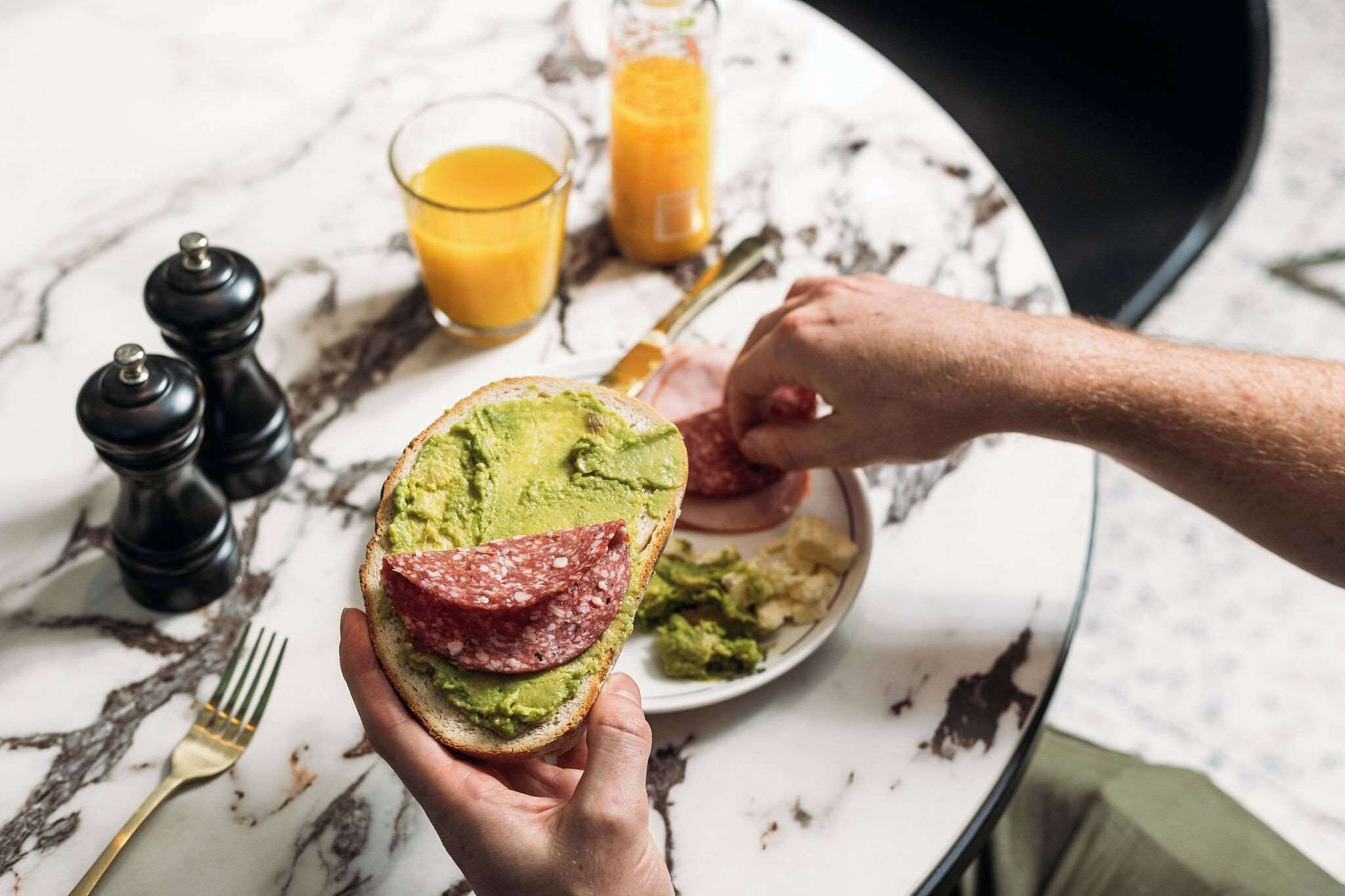 Iemand houdt een sandwich met avocado en salami vast op een marmeren tafel, met een glas sinaasappelsap en een bord chips.