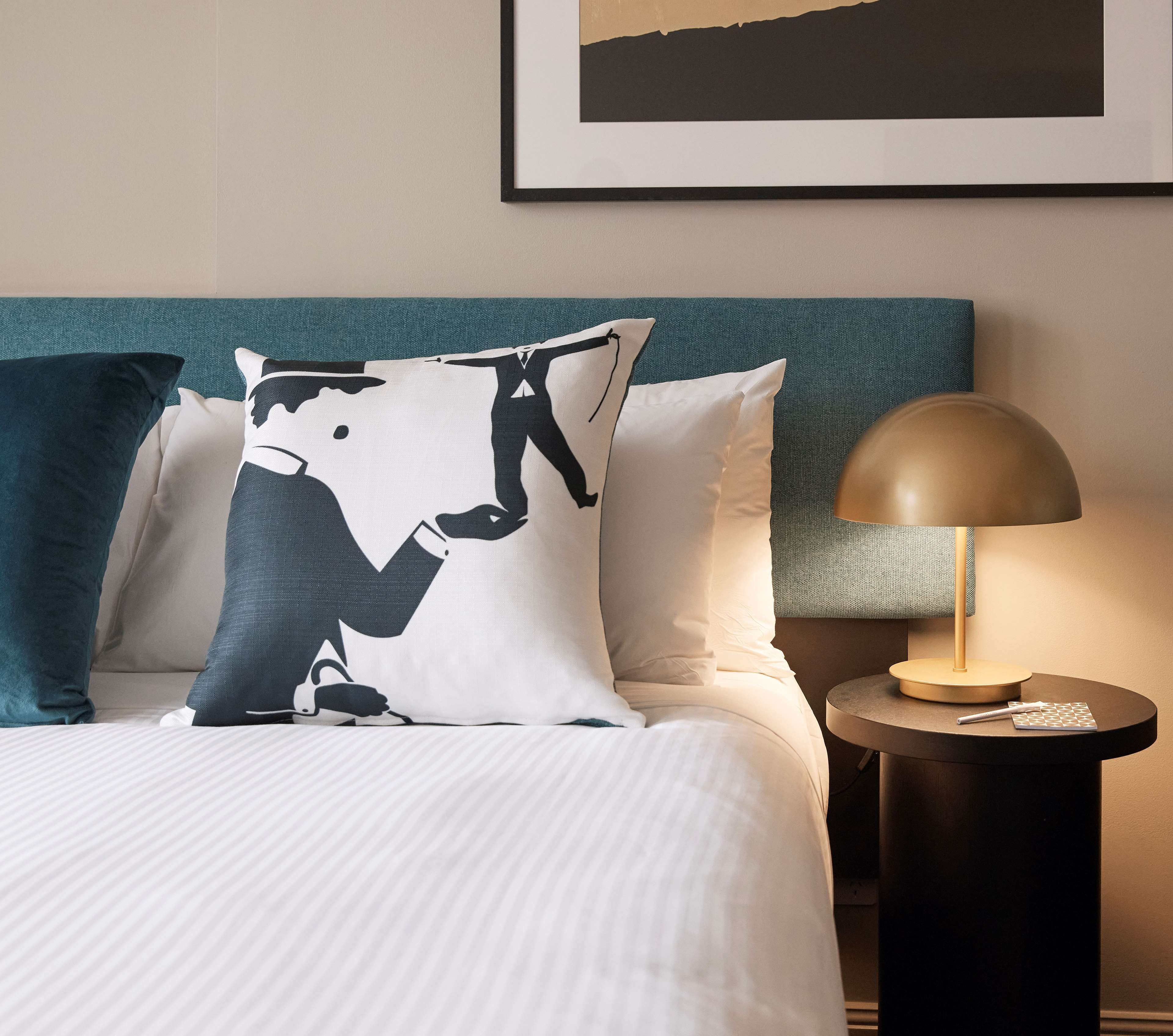 Un lit bien fait, avec une literie à rayures, une tête de lit turquoise et un coussin noir et blanc. Une table ronde avec une lampe et un stylo se trouve à côté du lit.