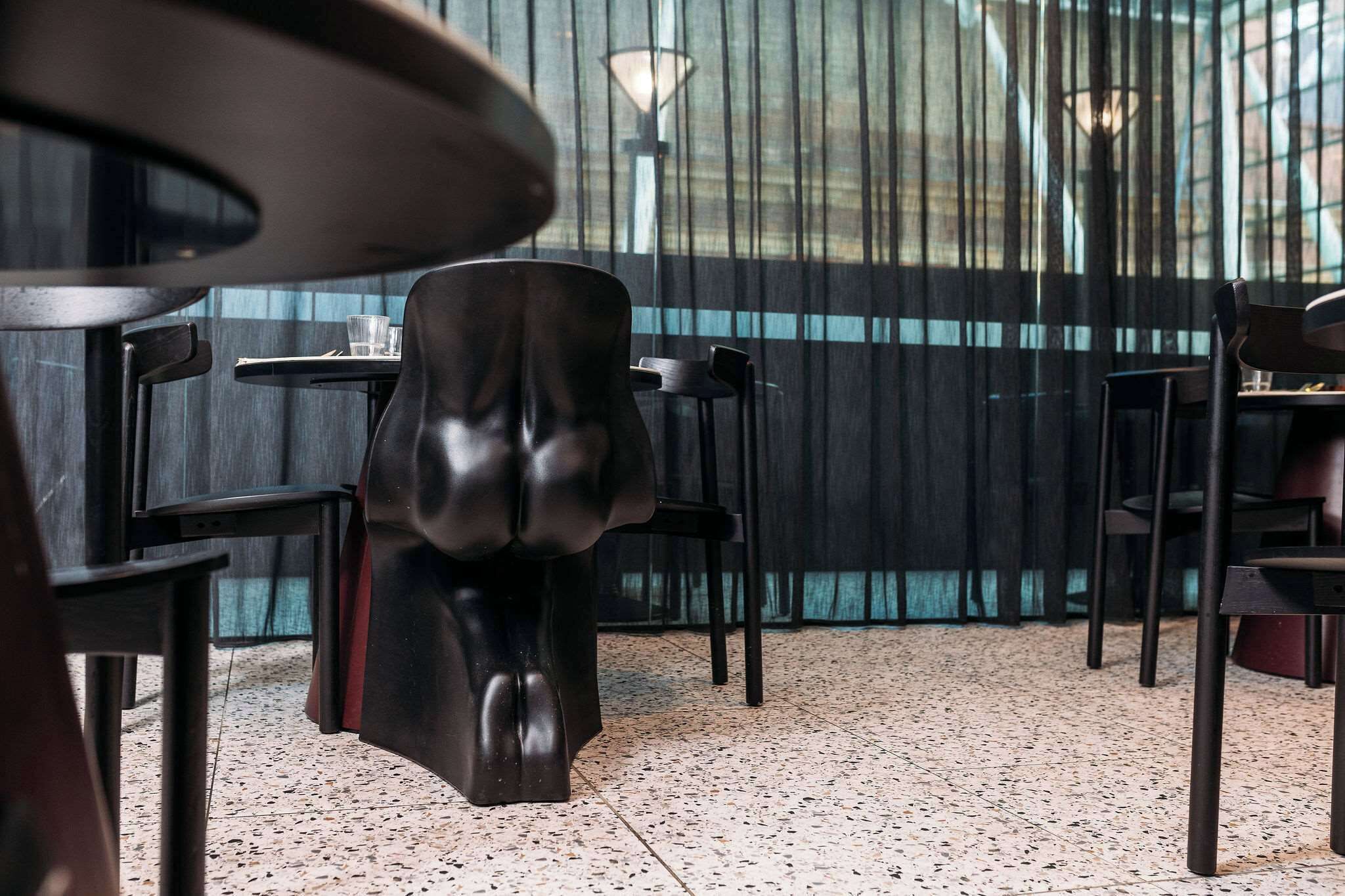 L’intérieur d’un restaurant avec des chaises et des tables noires, un sol à motifs et une vue à travers des rideaux transparents.