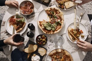 Ryhmä ihmisiä istuu marmoripöydän ääressä ja nauttii erilaisia ruokia, kuten pizzaa ja leipää.