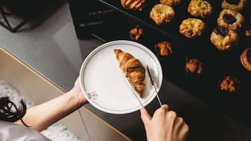A person holding a plate with a croissant and a knife, with a tray of pastries in the background.