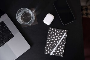 Una computadora portátil, un teléfono inteligente, un vaso de agua, un ratón blanco y una libreta con un diseño geométrico sobre una mesa de madera oscura.