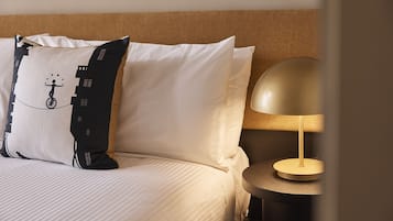 Una cama bien hecha con ropa blanca, un cojín blanco y negro con diseño de paisaje urbano y una mesa redonda con una lámpara dorada.