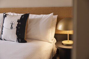 A neatly made bed with white linens, a black and white pillow with a cityscape design, and a round table with a gold lamp.
