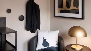 Una habitación moderna con un sillón negro, una mesa redonda con una lámpara, un cuadro abstracto enmarcado y una chaqueta negra colgada en la pared.