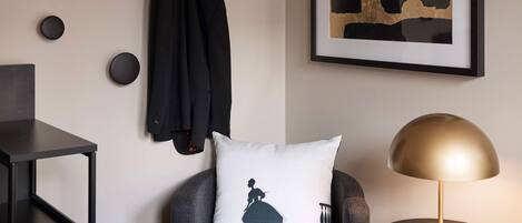 A modern room with a black armchair, a round table with a lamp, a framed abstract painting, and a black jacket hanging on the wall.