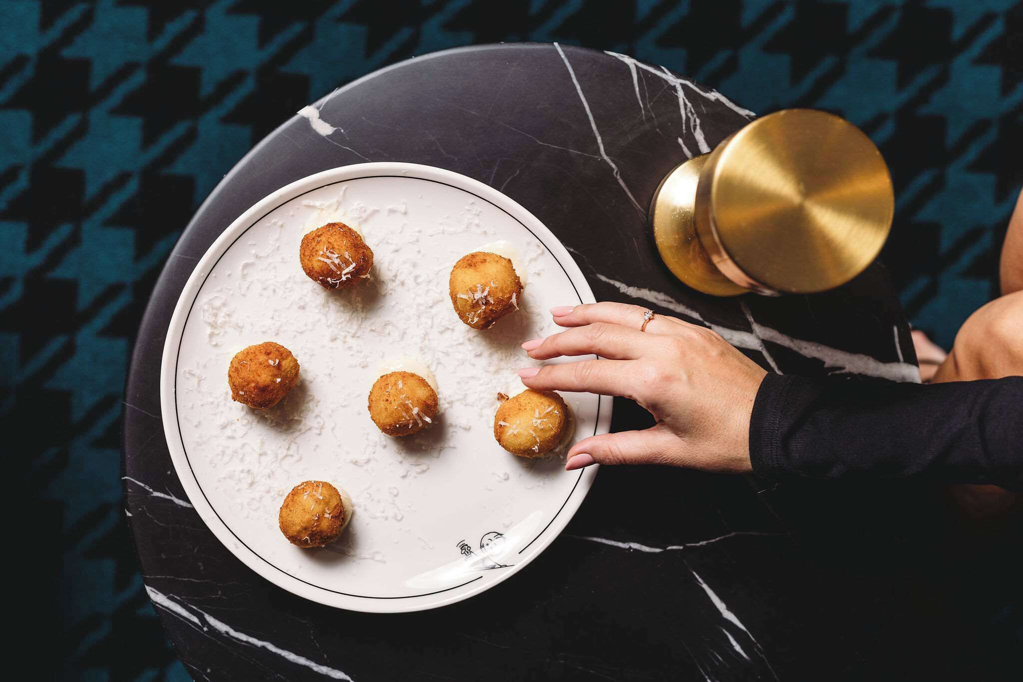 Une assiette blanche ornée d’un bord doré, sur laquelle sont disposés six aliments frits, de forme ronde et de couleur brun doré, le tout posé sur une surface en marbre foncé.