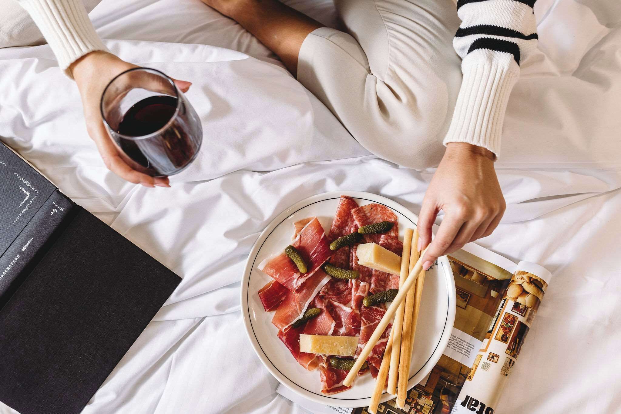 A person holding a glass of red wine and a plate of cured meats with pickles and breadsticks on a bed with white sheets.
