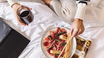 A person holding a glass of red wine and a plate of cured meats with pickles and breadsticks on a bed with white sheets.