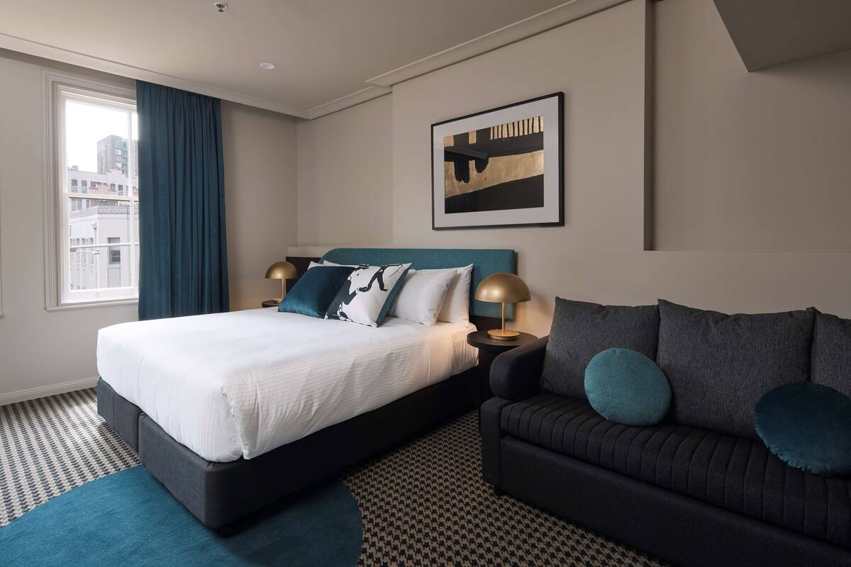 A modern hotel room with a large bed, a sofa, a framed artwork on the wall, and a view of the city through the window.