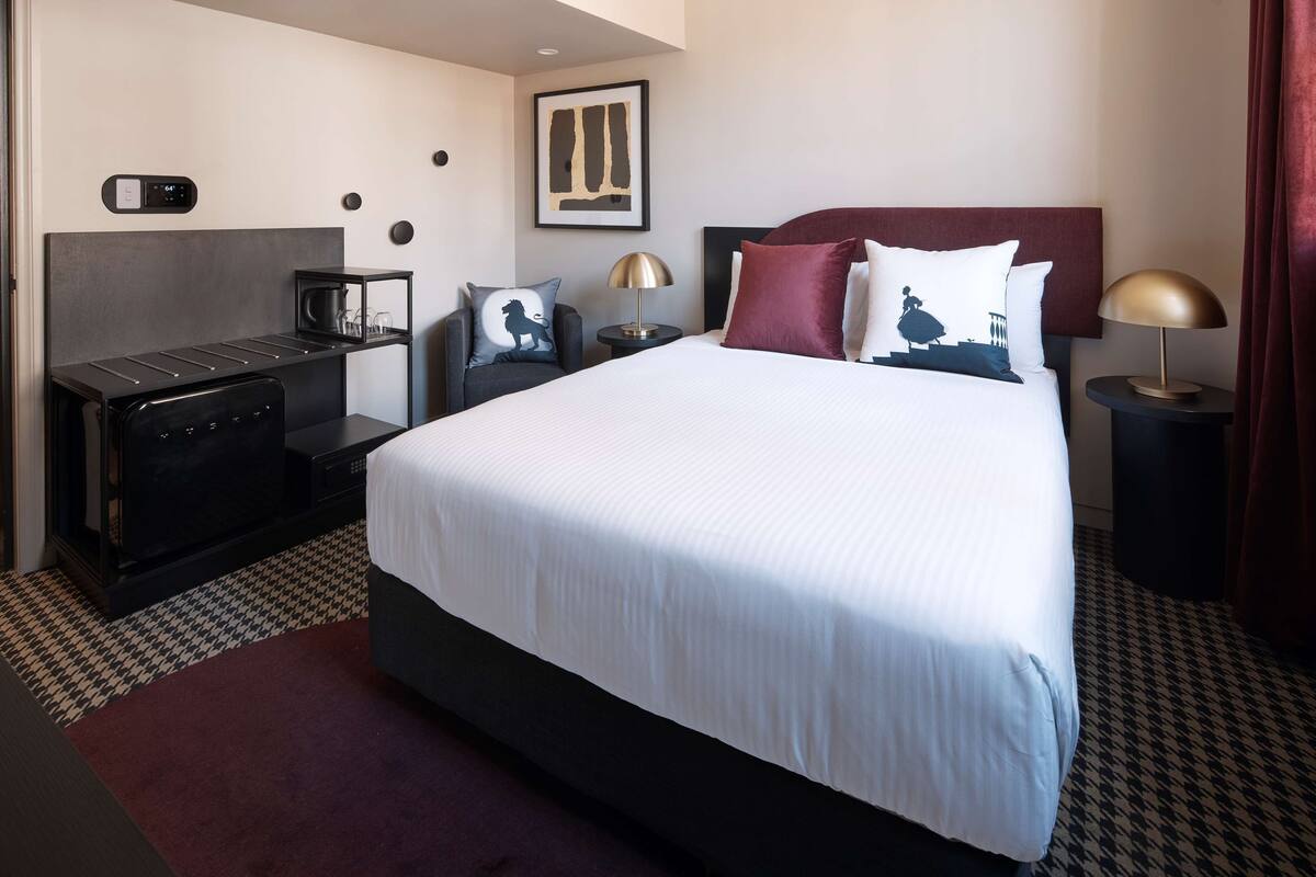 A hotel room with a large bed, a desk with a coffee machine, a TV, and a headboard with a red accent.
