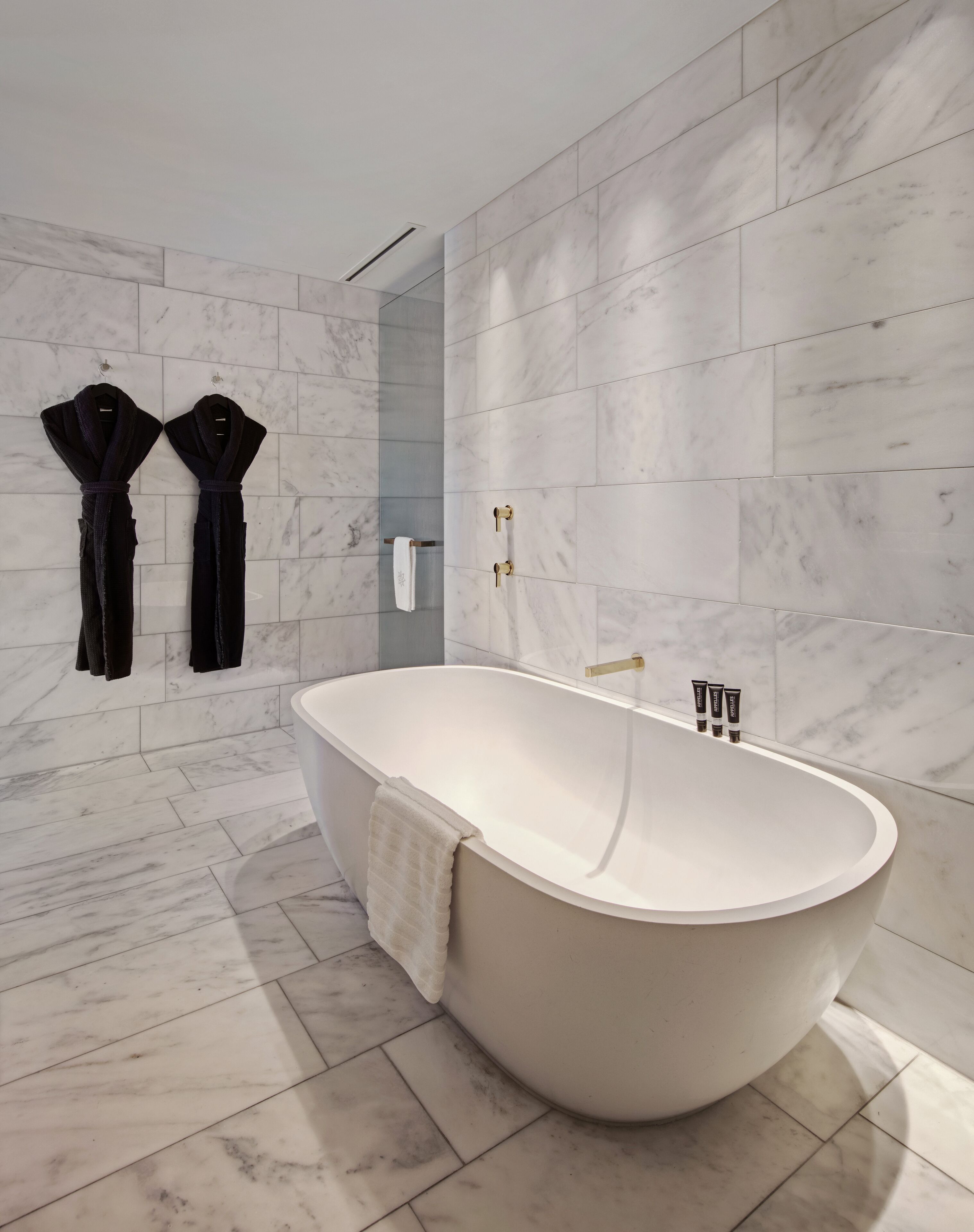 A modern bathroom with a freestanding bathtub, marble walls, and two black robes hanging on hooks.