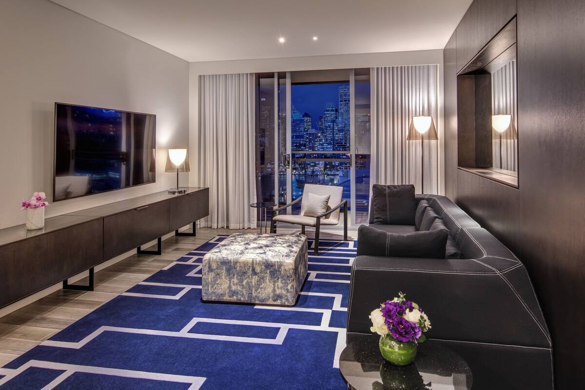 A modern hotel room with a large flat-screen TV, a sofa, a chair, and a view of the cityscape at night.