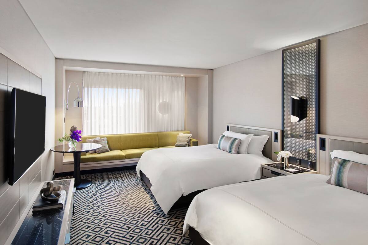 A hotel room with two beds, a sofa, a TV, a small table with a vase, and a patterned floor.