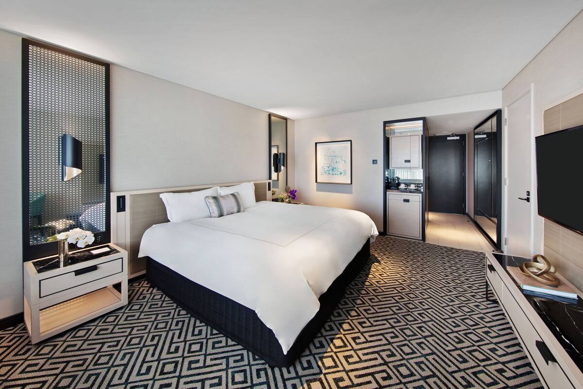 A modern hotel room with a large bed, a bedside table, a TV, and a bathroom visible through an open door.