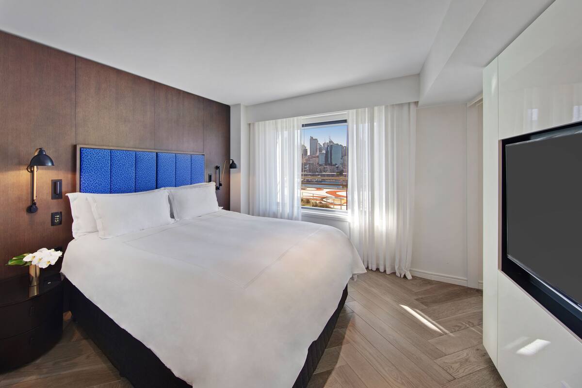 A hotel room with a large bed, a TV, a nightstand with a lamp, and a window with a city view.