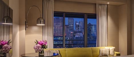 Habitación de hotel con una cama, un sofá verde, una mesita redonda con un florero y un espejo, y vistas a un paisaje urbano nocturno.