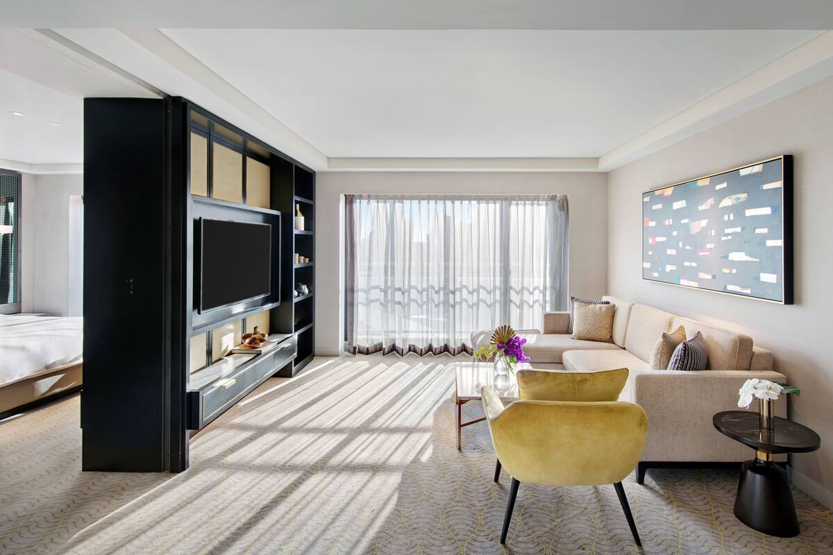 A modern hotel room with a large flat-screen TV, a sofa, a yellow armchair, a small table, and a large window with sheer curtains.