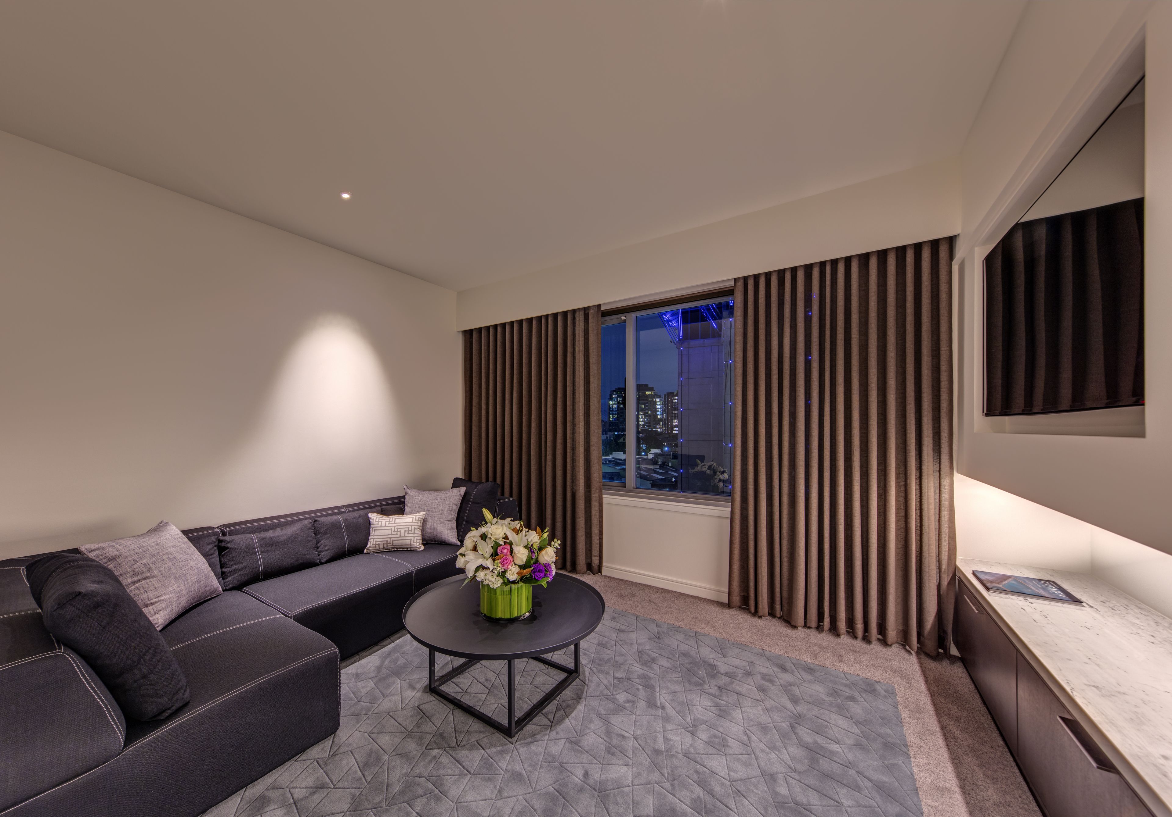 A modern living room with a sofa, a coffee table with flowers, a TV, and a window with curtains.