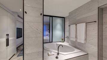 A modern bathroom with a large freestanding bathtub, a glass shower enclosure, and a visible bedroom with a bed and curtains.