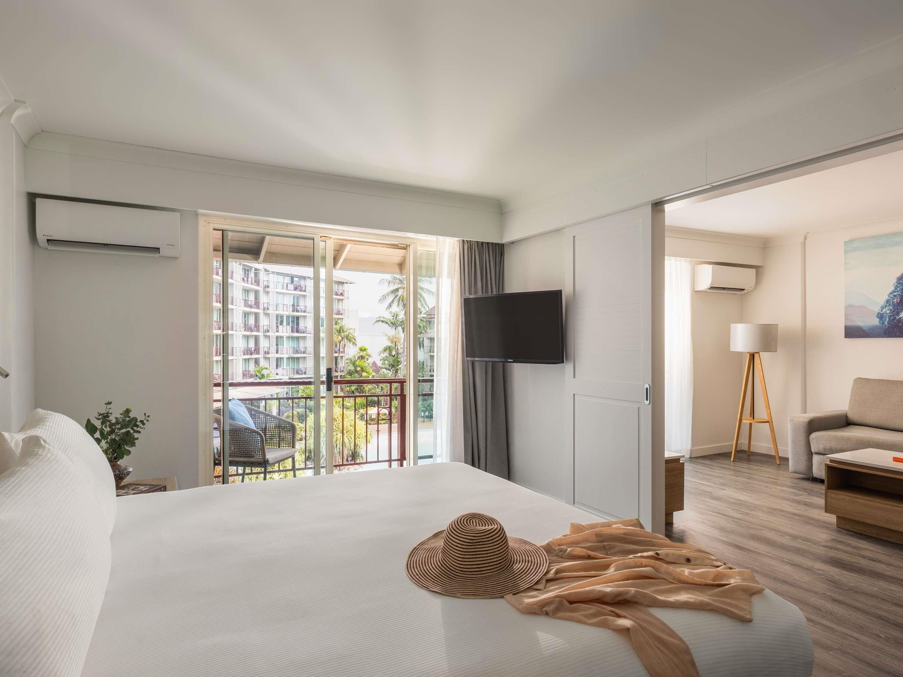 A hotel room with a bed, a TV, a balcony with a view of buildings, and a desk with a lamp.