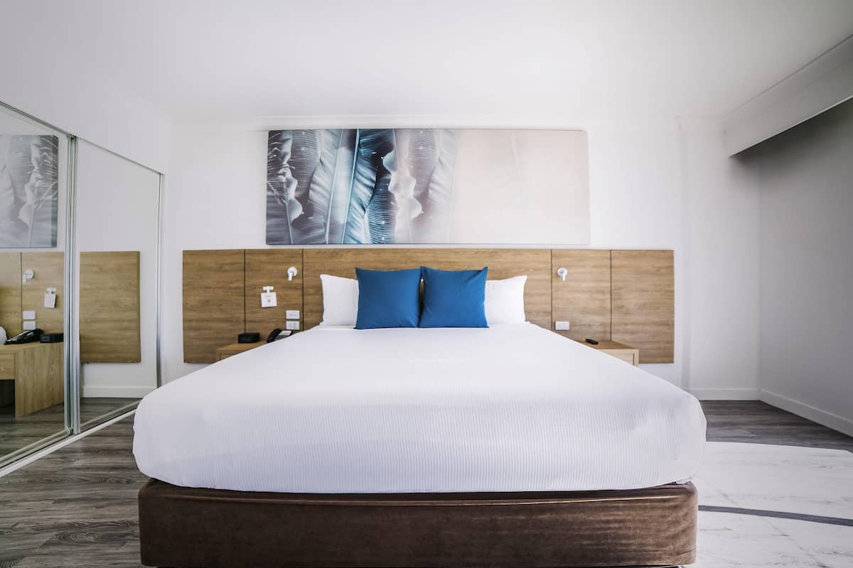 A modern hotel room with a large bed, wooden headboard, bedside tables, and a large mirror.
