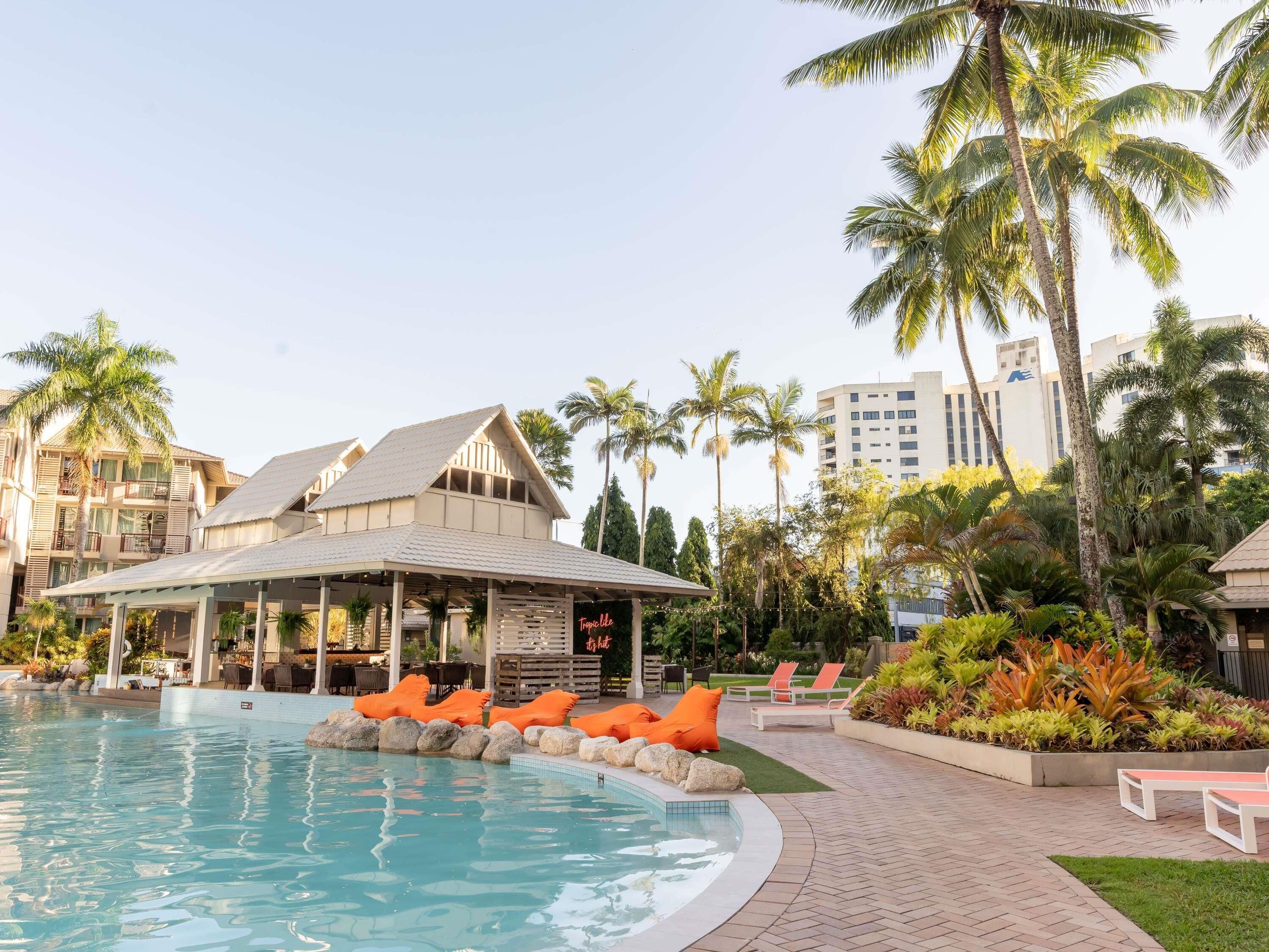 A swimming pool with lounge chairs and a covered area, surrounded by palm trees and a building with balconies.