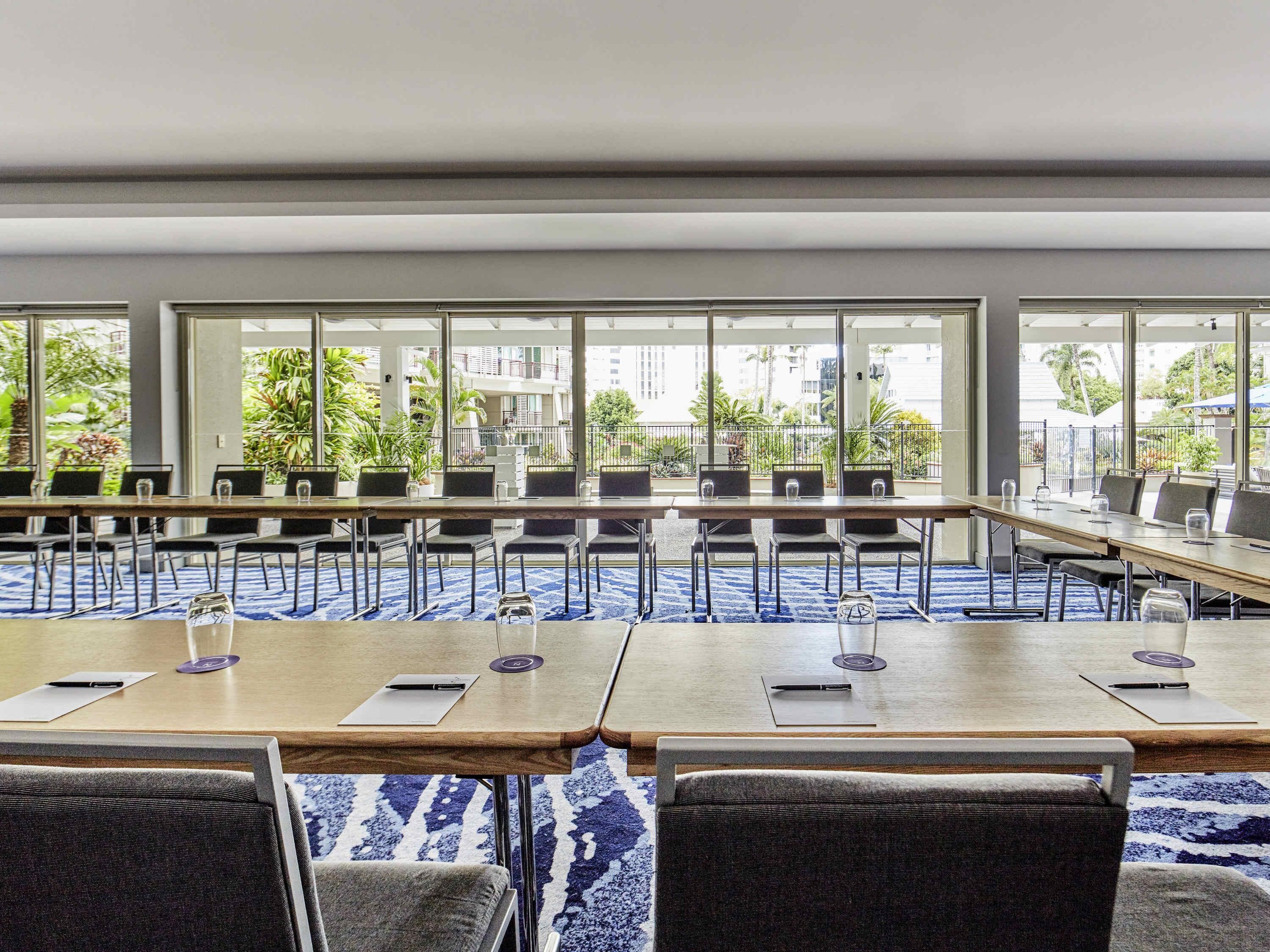 Un salón de conferencias con mesas largas, sillas y ventanas amplias que ofrecen vistas a zonas verdes y edificios.
