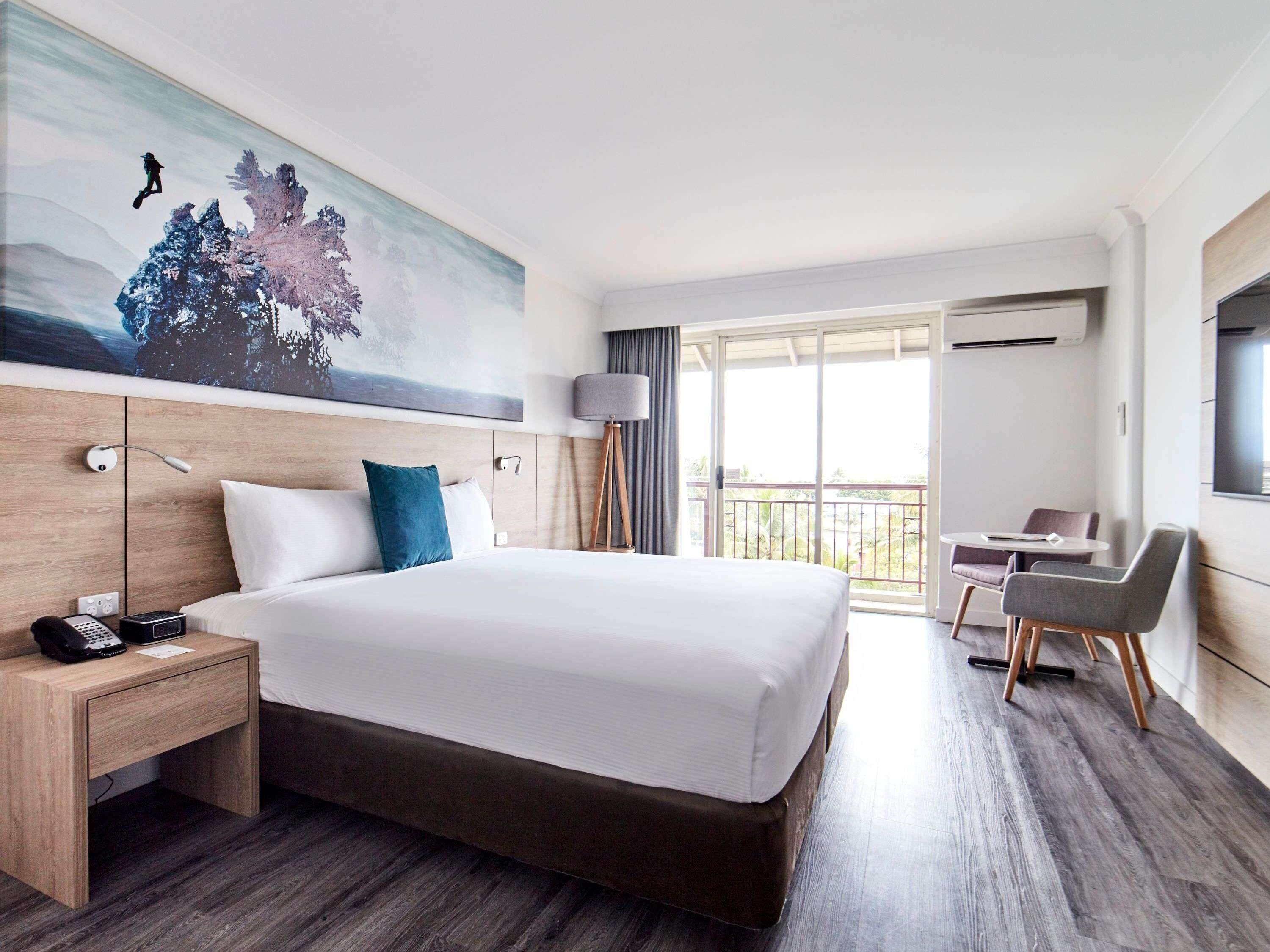 A hotel room with a large bed, a desk with a phone, a chair, a balcony with a view, and a painting on the wall.