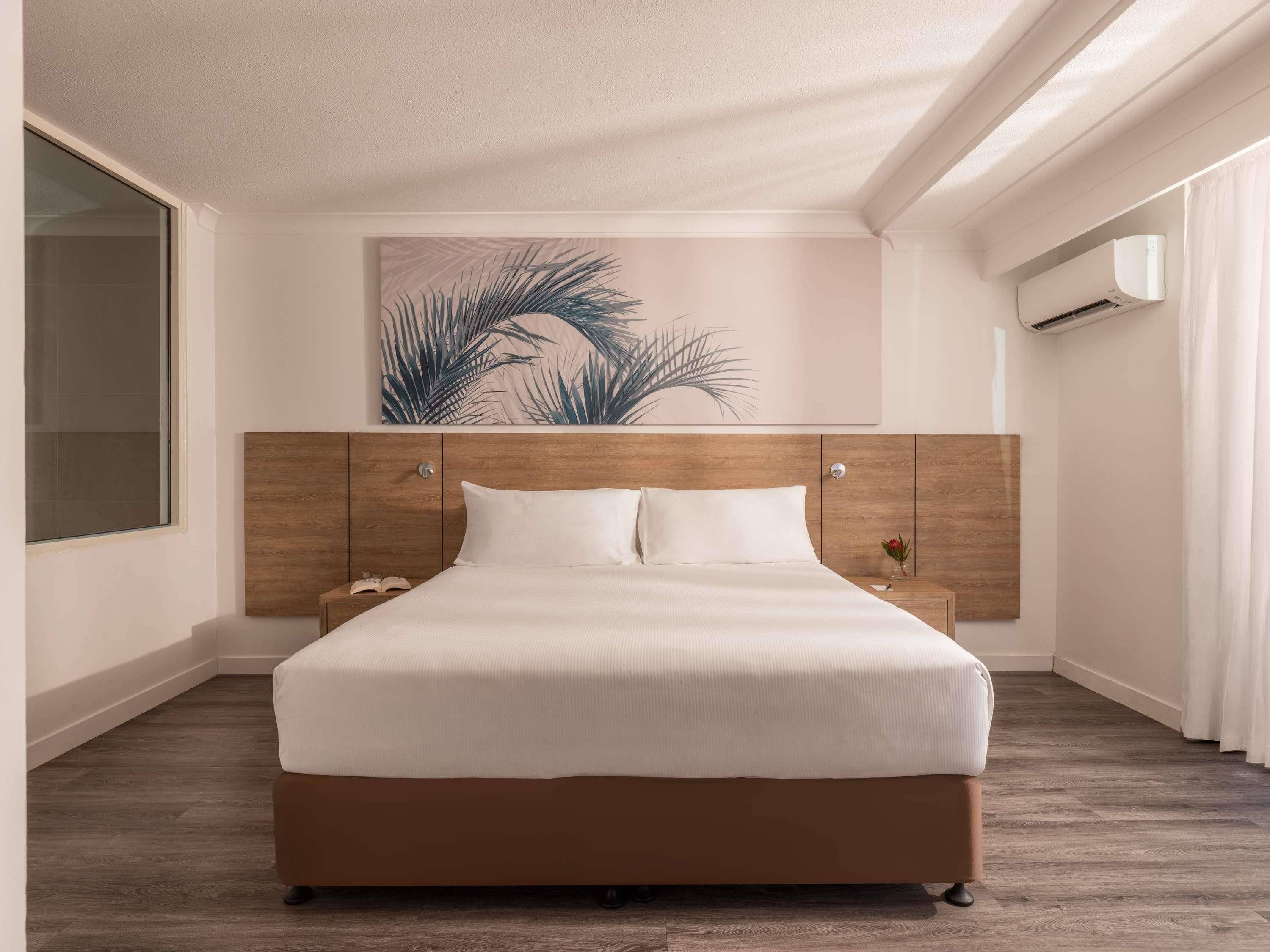 Habitación de hotel con cabecera de madera, cama grande con ropa blanca, mesita de noche con lámpara y unidad de aire acondicionado fijada a la pared.