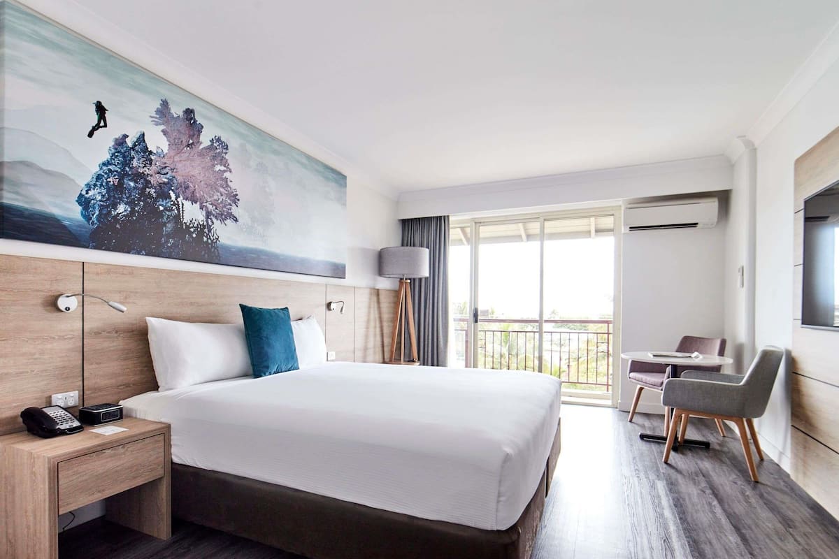 A hotel room with a large bed, a desk with a phone, a chair, a balcony with a view, and a painting on the wall.