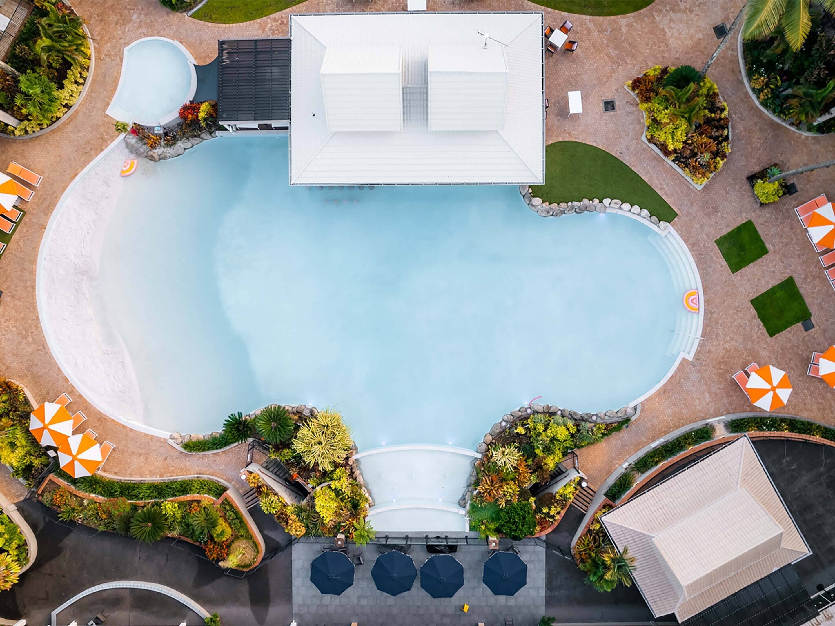 An aerial view of a hotel with a central swimming pool, surrounded by lounge areas with umbrellas, and a covered seating area.