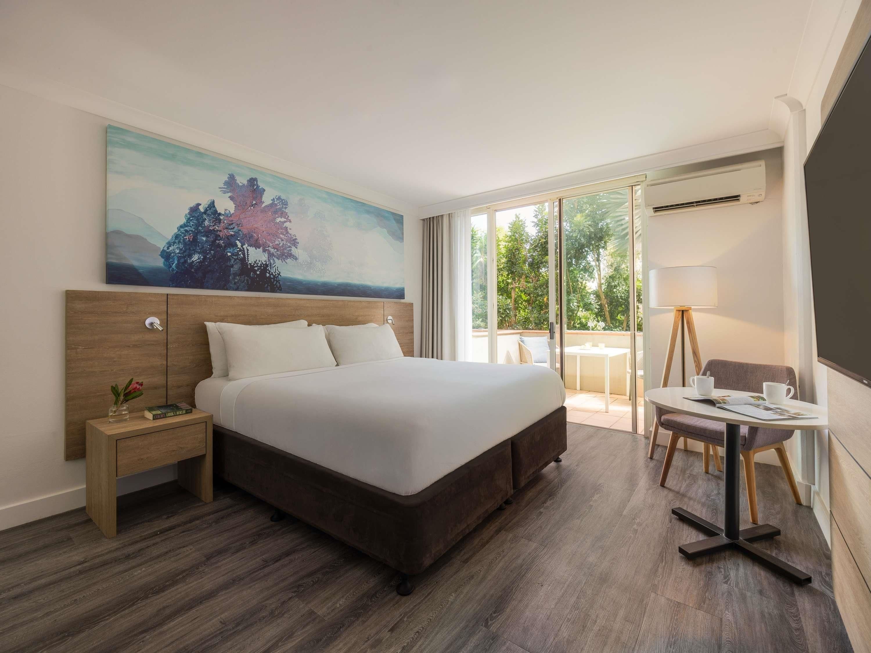 Une chambre d’hôtel dotée d’un grand lit, d’une table de chevet, d’un bureau avec une chaise et offrant une vue sur l’extérieur grâce à une porte-fenêtre coulissante.
