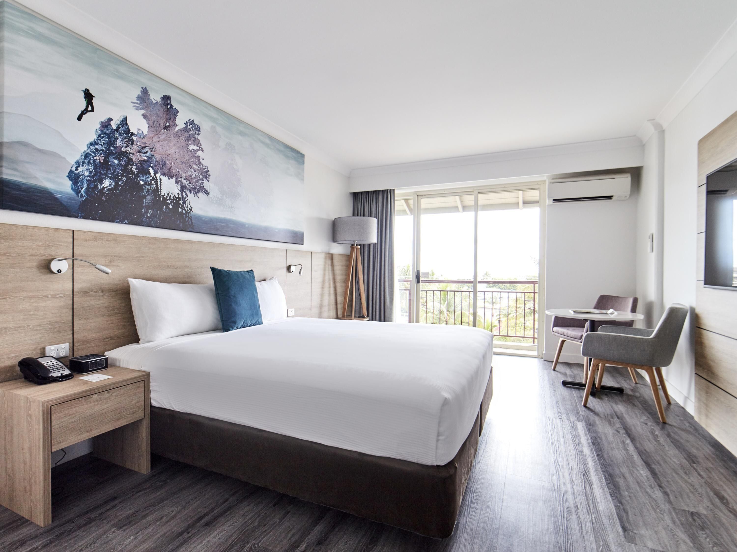 A hotel room with a large bed, a desk with a chair, a balcony with a view, and a painting on the wall.