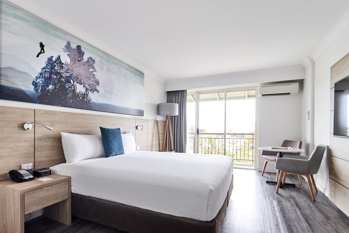 A hotel room with a large bed, a desk with a chair, a balcony with a view, and a painting on the wall.