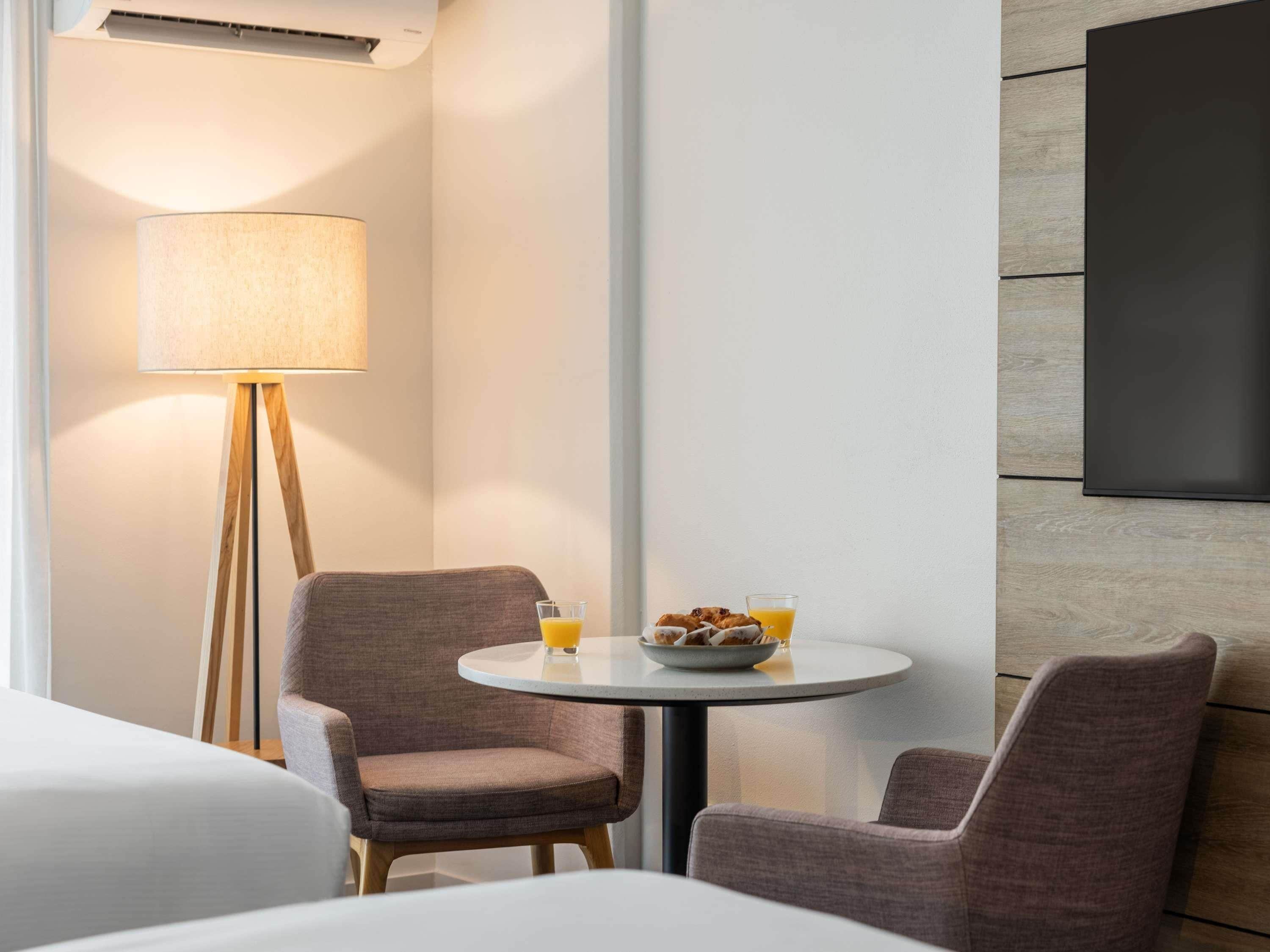 A hotel room with a round table, two chairs, a lamp, and a flat-screen TV mounted on the wall.