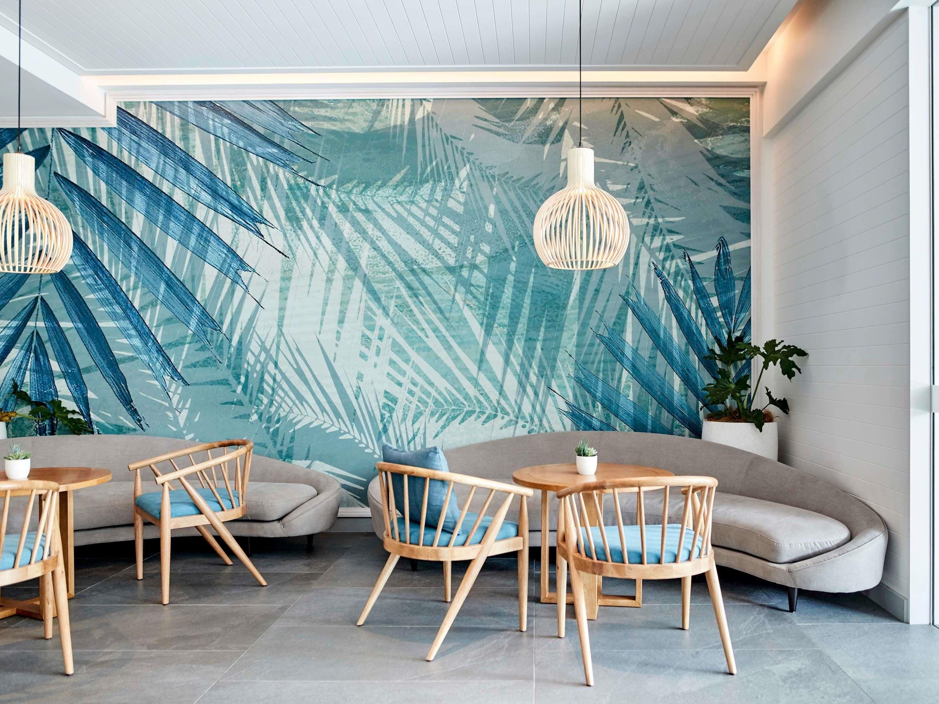 Um interior moderno com uma grande parede estampada com folhas, cadeiras de madeira, um sofá e luminárias pendentes.