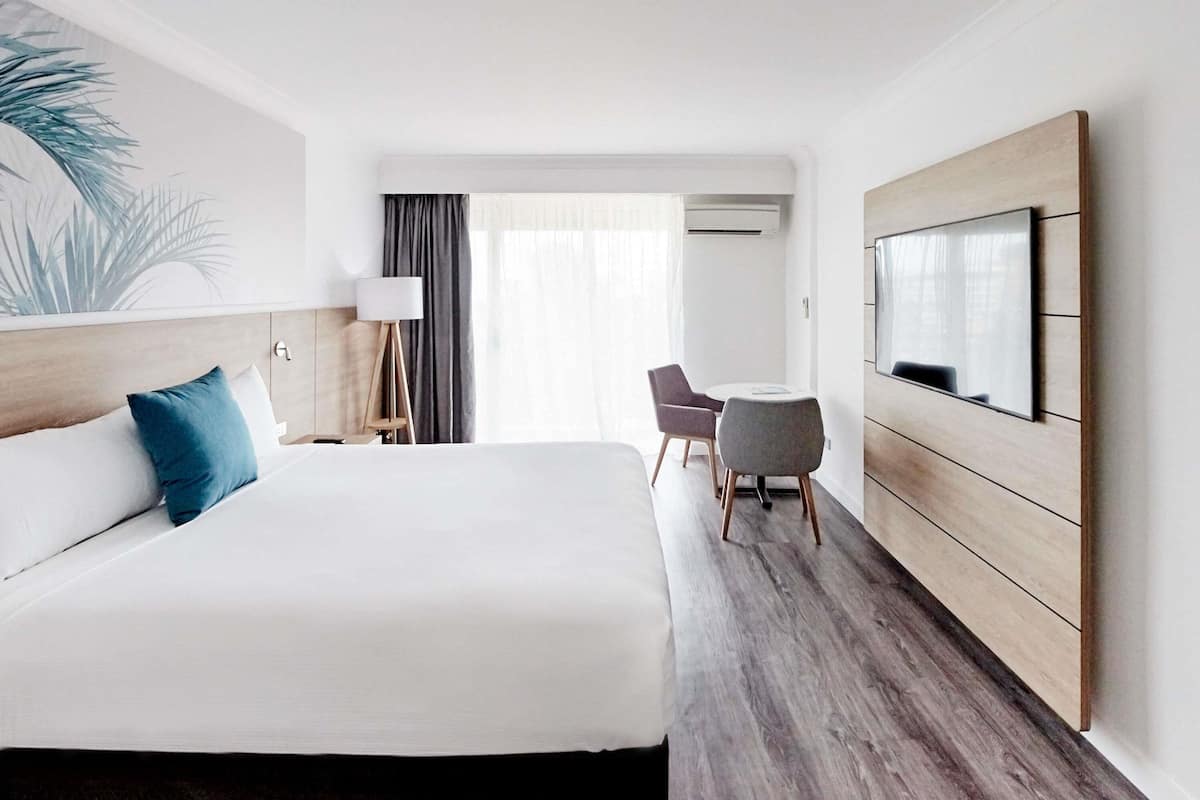 A hotel room with a large bed, a desk with two chairs, a TV mounted on the wall, and a window with curtains.