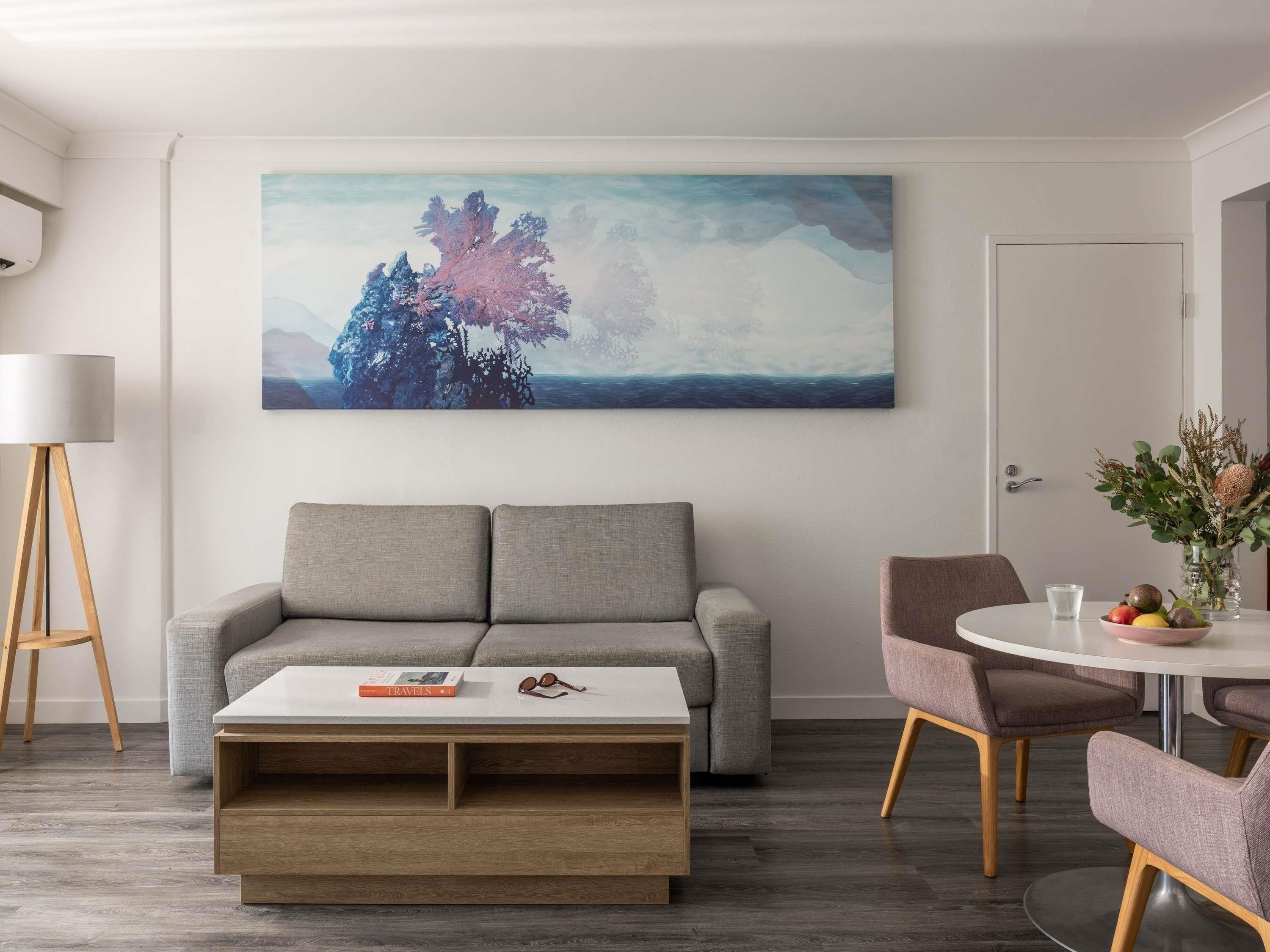 En moderne stue med en grå sofa, et sofabord, et billede af et træ, en gulvlampe og et spiseområde med stole og et bord.