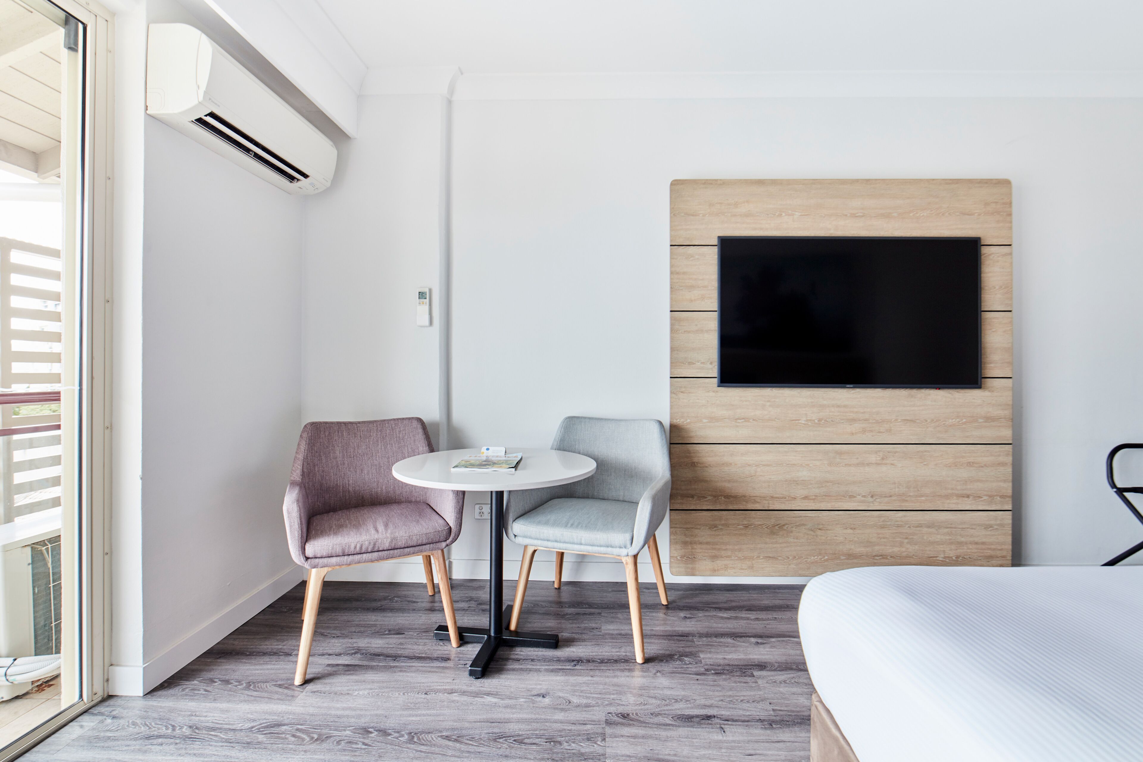 A modern hotel room with a bed, a TV mounted on a wooden panel wall, a small round table with two chairs, and an air conditioning unit.
