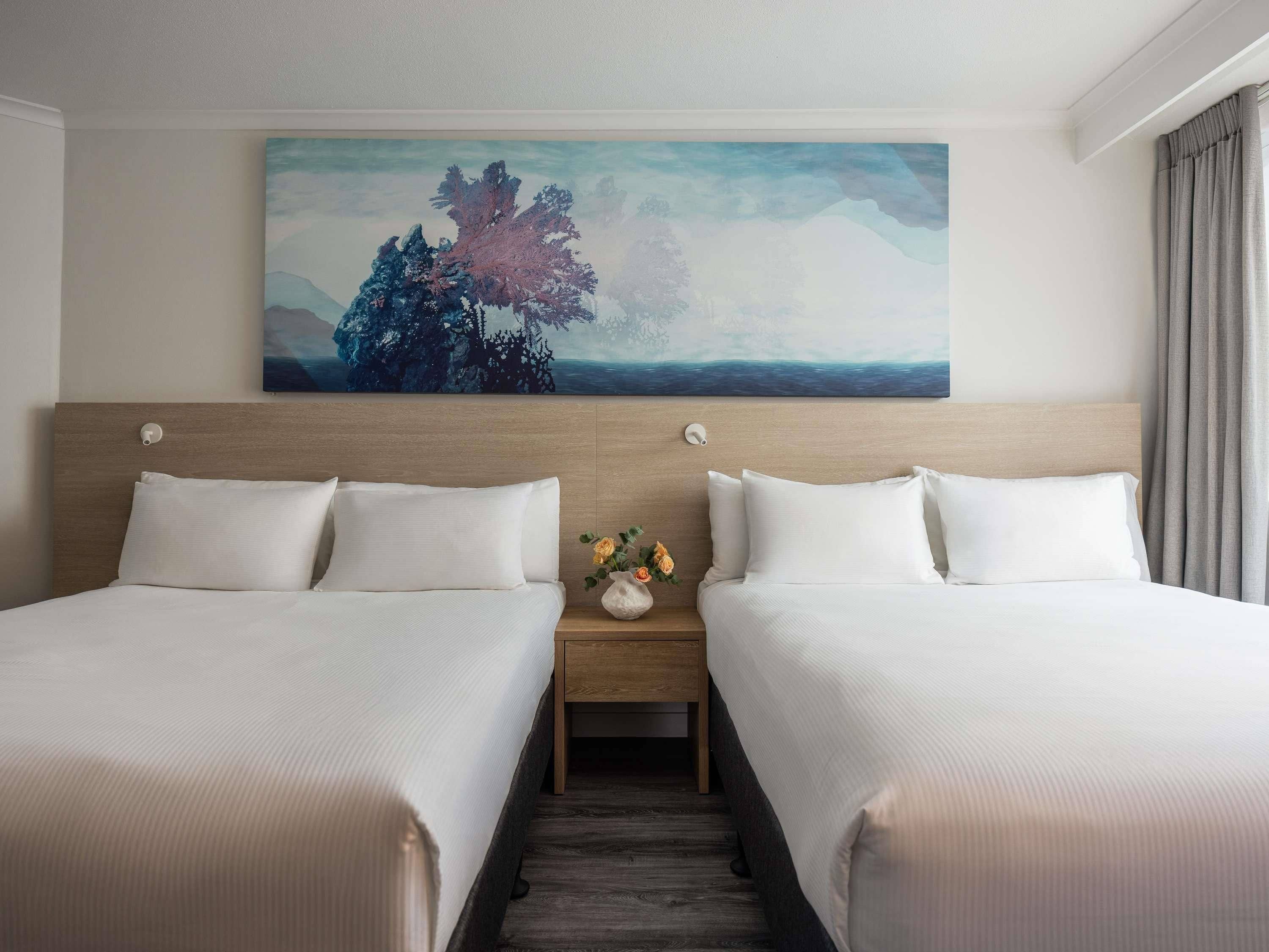 A hotel room with two beds, a nightstand with a vase of flowers, and a painting on the wall.