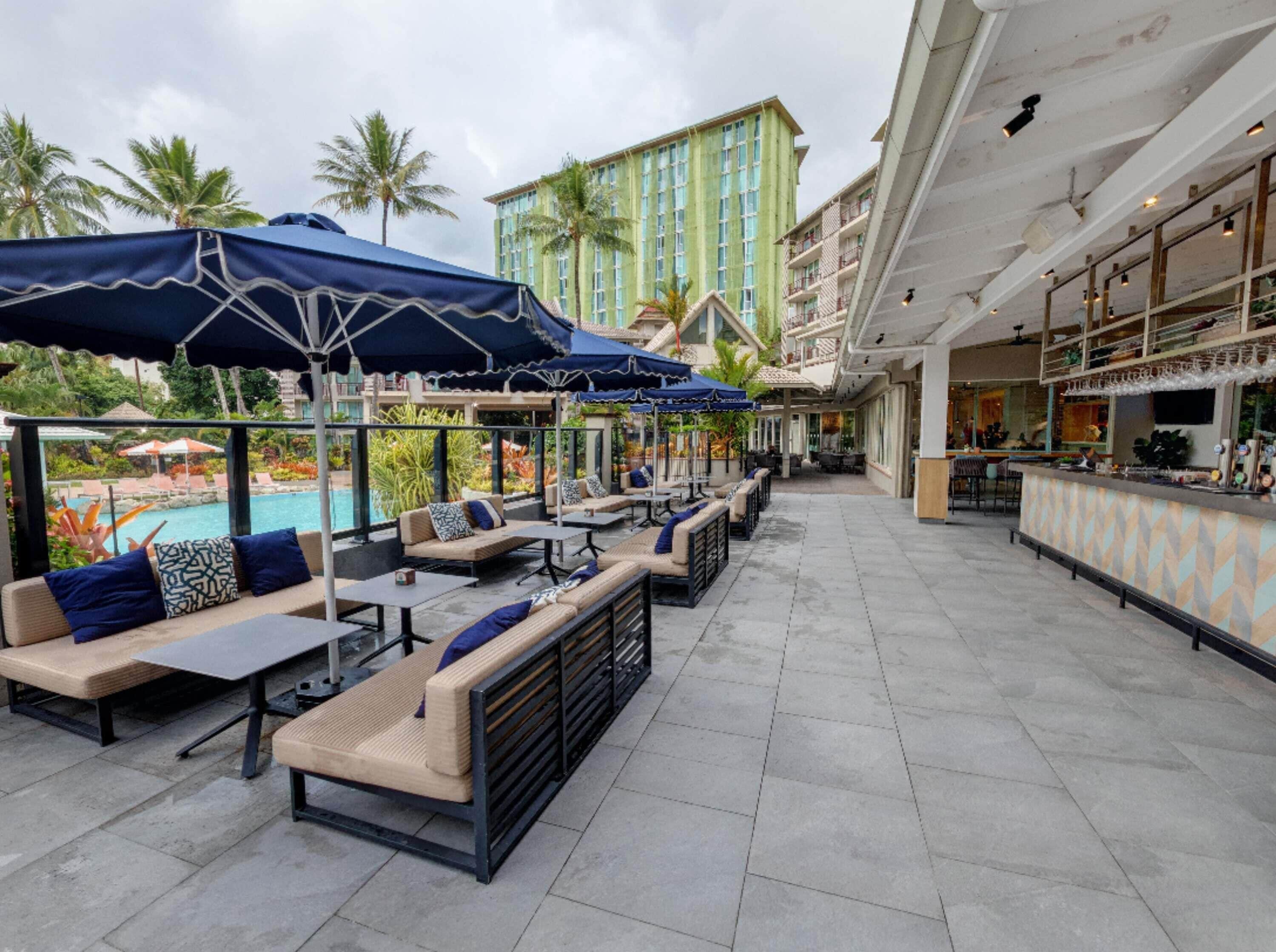 A rooftop bar with outdoor seating, a pool, and a view of palm trees and a hotel building.