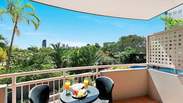 A balcony with a table and chairs, a plate of fruit and two glasses of orange juice, and a cityscape view in the background.