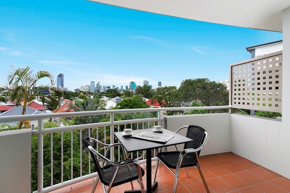 Un balcon avec une table et des chaises, donnant sur un paysage urbain avec des bâtiments et de la verdure.
