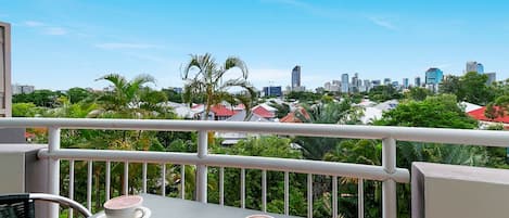 A balcony with two tables, each with a cup of coffee, overlooking a cityscape with buildings and greenery.